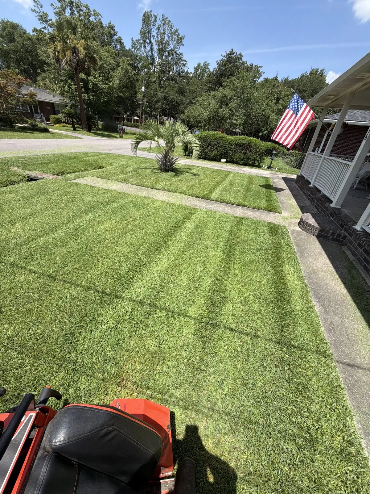 A zero-turn mower's view across a freshly striped front lawn with an American flag on the porch