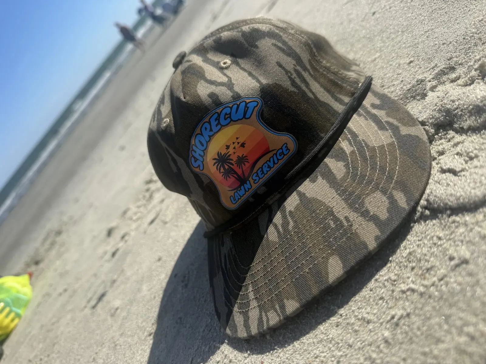 A camo ShoreCut Lawn Service hat on the beach at Isle of Palms